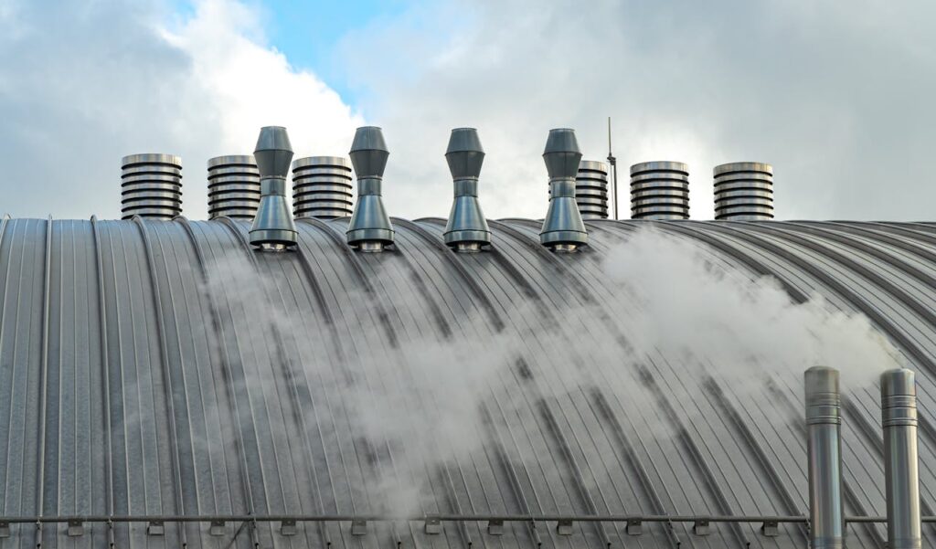 Photo of industrial rooftop with metal vents and steam against a cloudy sky.